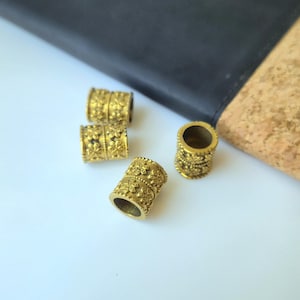 May include: Four cylindrical metal beads with a brass finish, each featuring a repeating embossed floral pattern. The beads are hollow with a central opening, displaying a textured surface with small flower motifs arranged in horizontal bands.