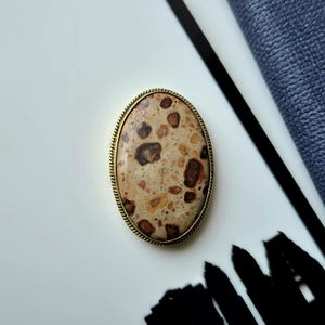 May include: An oval brooch featuring a polished, speckled stone with a beige base and dark brown, tan, and cream-colored inclusions. The stone is encased in a thin, textured gold-toned metal frame with a rope-like edge detail around the perimeter.