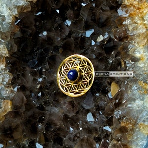 May include: A gold-toned metal pendant featuring a flower of life design with a blue gemstone in the center. The pendant is on a bed of clear quartz crystals.