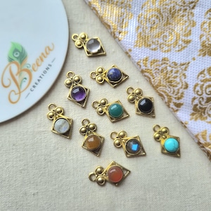 May include: Ten gold-toned metal jewelry charms, each featuring a diamond-shaped frame with a circular gemstone in various colors including purple, blue, green, black, white, orange, and turquoise, with a small loop at the top for attachment.