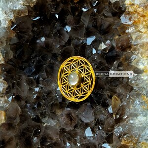 May include: A gold-toned metal pendant featuring a flower of life design with a white stone in the center. The pendant is resting on a bed of dark gray crystals.