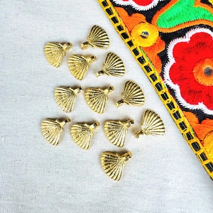 Puede incluir: Una colección de pequeños adornos decorativos dorados, cada uno con forma de abanico estilizado. Los adornos están dispersos sobre una tela de color claro, con una porción de un textil bordado colorido en el fondo.