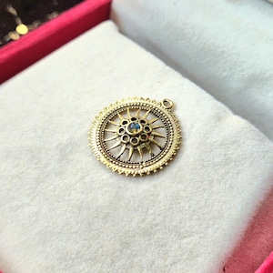 Puede incluir: Un colgante dorado con un diseño de sol. El colgante presenta una piedra azul central y está rodeado de detalles intrincados. El colgante es redondo y tiene un pequeño lazo en la parte superior para una cadena. El colgante se muestra sobre una superficie blanca.