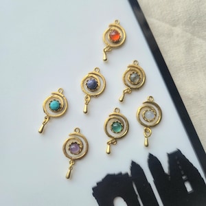 Op de afbeelding: Zeven goudkleurige metalen bedels, elk met een spiraalontwerp en een kleine bungelende druppel. Elke bedel is bezet met een cabochonsteen in een andere kleur, waaronder tinten oranje, blauw, paars, grijs, turkoois, groen en wit.