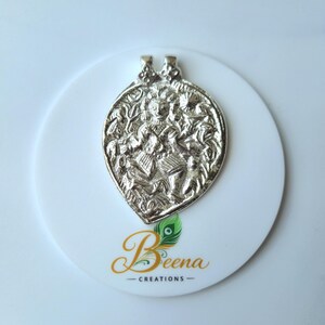 May include: Silver-colored pendant with an intricate design, possibly depicting a deity or figure. The pendant is leaf-shaped and rests on a white circular surface. The logo "Beena Creations" is visible.