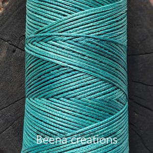 Puede incluir: Un primer plano de un carrete de cordón de algodón encerado azul turquesa. El cordón está enrollado apretado y tiene un acabado liso y pulido. El texto "Beena creations" es visible en el carrete.