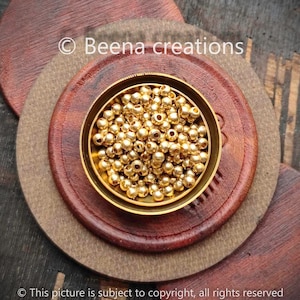 以下が含まれることがあります： 金色の丸いビーズでいっぱいになった小さなボウルをクローズアップした写真。ボウルは木製の表面の上に置かれています。