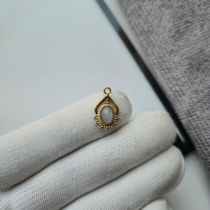 May include: A small gold-toned metal pendant featuring an oval-shaped, translucent white stone set in the center. The pendant has a decorative, textured border with small bead-like accents and a single jump ring at the top for attachment.