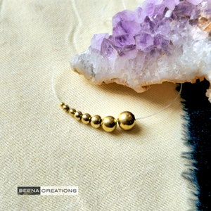 Könnte beinhalten: Eine goldene Halskette mit einer abgestuften Kette aus runden Perlen. Die Halskette wird auf einer weißen Oberfläche mit einem violetten Amethystkristall im Hintergrund gezeigt.