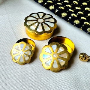 Può includere: Un set di tre piccole scatole in metallo color oro con coperchi a forma di fiore intarsiati con madreperla. Le scatole sono disposte su una superficie chiara, con un tessuto blu scuro con dettagli dorati sullo sfondo.