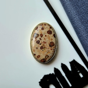 Puede incluir: Un broche de forma ovalada que presenta una piedra pulida con un patrón moteado de tonos beige, crema y marrón oscuro. La piedra está engastada en un marco de metal dorado fino y texturizado con un borde trenzado de aspecto clásico.