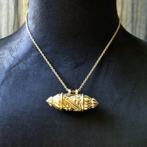 Puede incluir: Collar con colgante dorado. El colgante tiene forma de cápsula alargada y ornamentada con intrincados detalles florales y de volutas. La cadena es de estilo eslabón delicado. El collar se exhibe en un maniquí negro.