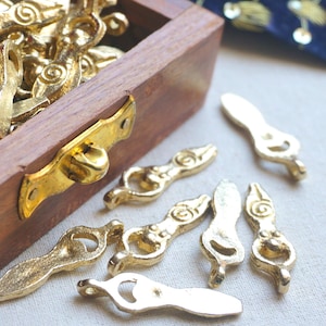 Puede incluir: Primer plano de varias piezas decorativas de metal dorado, algunas dispersas y otras dentro de una caja de madera. Las piezas tienen un diseño único y estilizado con líneas curvas y un lazo en un extremo. La caja tiene un cierre dorado.