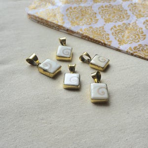 May include: Five square pendants featuring a white shiva eye shell inlay with a natural spiral pattern, each set in a polished gold-tone metal frame with a top bail for hanging. The pendants are arranged in a cluster on a neutral fabric surface.