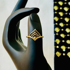 Op de afbeelding: Een gouden ring met een geometrisch ontwerp met een centrale cirkel en ingewikkelde details. De ring is op een zwart handmodel.