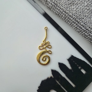 Op de afbeelding: Een goudkleurige metalen hanger met een ingewikkeld, wervelend unalome-ontwerp. De bedel heeft een kleine ronde lus aan de bovenkant voor bevestiging, met een gladde, gepolijste afwerking die het licht reflecteert op de gebogen, verbonden lijnen.
