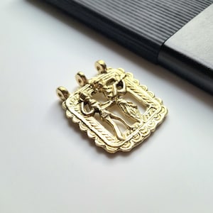 May include: A gold-colored pendant with a rectangular frame and a detailed relief of two figures. The pendant has three loops for hanging and a decorative, scalloped edge. The background is white, with a black object in the upper right corner.