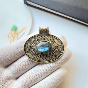 May include: An oval-shaped pendant featuring a central iridescent blue labradorite gemstone set in an antique gold-toned metal frame. The metalwork displays intricate filigree patterns, beaded borders, and a small decorative bail at the top.