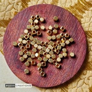 Peut inclure: Gros plan sur un tas de perles en métal de couleur dorée sur une surface en bois. Les perles sont de forme carrée avec un bord légèrement arrondi. Les perles sont disposées de manière aléatoire.