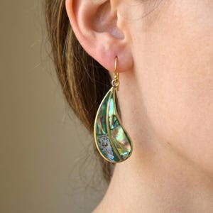 May include: A teardrop-shaped dangle earring featuring iridescent abalone shell inlays set within a gold-toned metal frame. The shell displays a vibrant mix of green, blue, and pearlescent tones, secured by a simple gold-toned hook wire.
