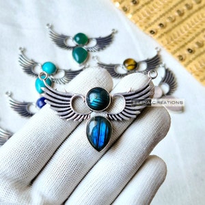 Op de afbeelding: Zilveren hanger met twee blauwe labradorite stenen ingelegd in een vleugelontwerp. De hanger ligt op een wit oppervlak. De tekst "BEENA CREATIONS" is zichtbaar op het oppervlak.