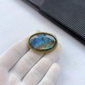 May include: An oval brooch featuring a polished labradorite gemstone with iridescent blue and gold flashes. The stone is set within a textured, twisted gold-tone metal frame that follows the oval shape of the cabochon, creating a decorative border.