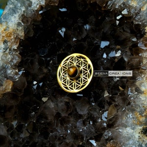 May include: Gold-toned metal flower of life design with a tiger's eye stone in the center. "BEENA CREATIONS" is written in the bottom right corner of the image.