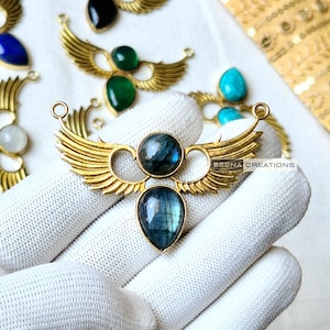 May include: Gold tone pendant with two teardrop-shaped labradorite stones and a pair of wings. The pendant is on a white surface. "BEENA CREATIONS" is printed on the pendant.