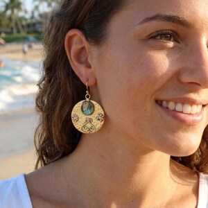 May include: A gold-toned circular drop earring featuring a hammered texture, a small central iridescent blue and green inlay, and decorative embossed scrollwork patterns. The earring hangs from a simple gold-toned hook wire attachment.