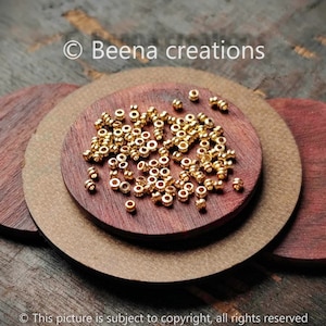 May include: A close-up of a pile of gold-coloured metal beads on a wooden surface. The beads are small and round, with a hole in the centre. They are arranged in a random pattern.