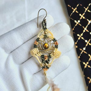 May include: A gold-toned lotus flower charm with a tiger's eye stone in the centre, surrounded by a macrame design in cream, green, and brown threads. The charm is attached to a gold-toned hook and has a feather tassel.