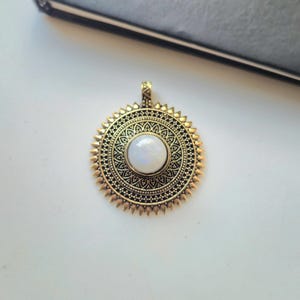 Op de afbeelding: Een ronde hanger met een antiek goudkleurig metalen frame met ingewikkelde filigraanpatronen en een stekelige buitenrand. Een gladde, ronde, iriserende witte cabochonsteen is in het midden van het medaillon gezet, dat een klein oogje heeft.