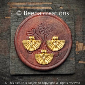 Op de afbeelding: Drie gouden metalen hangers met een halve maan vorm en ingewikkelde ontwerpen. De hangers liggen op een bruin houten oppervlak.