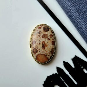 Puede incluir: Un broche de forma ovalada que presenta una piedra pulida y moteada con inclusiones en tonos beige, canela y marrón oscuro. La piedra está engastada en un marco de metal dorado fino y texturizado con un delicado patrón de cuerda en el borde.