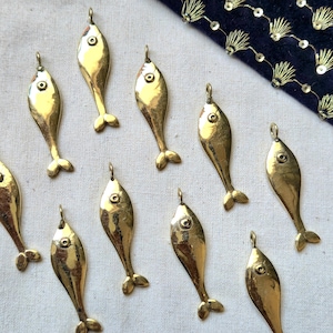 Può includere: Una collezione di piccoli ciondoli a forma di pesce color oro. Ogni ciondolo ha un anello nella parte superiore per appenderlo, un occhio rotondo e una piccola pinna caudale. I ciondoli sono disposti su una superficie chiara.