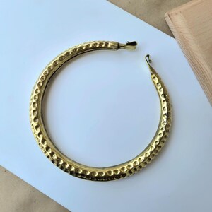 Puede incluir: Un collar rígido circular de latón con una textura martillada y con hoyuelos en toda su superficie. La pieza está abierta en la parte superior, con cada extremo terminando en un pequeño detalle de gancho curvado. El metal tiene un acabado dorado.