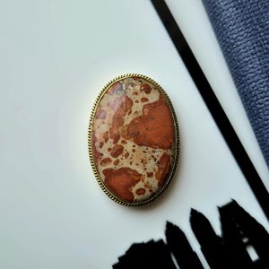 Puede incluir: Un broche de forma ovalada que presenta una piedra pulida con un patrón moteado de colores rojo tierra, beige y crema. La piedra está engastada en un marco de metal fino y texturizado en tono dorado que sigue el contorno ovalado.