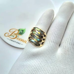 May include: A gold-toned ring featuring a wide, open-work band with wavy, horizontal cutouts. Inlaid within the metal frame are iridescent, multi-colored abalone shell segments that display shifting hues of green, blue, and purple in the light.