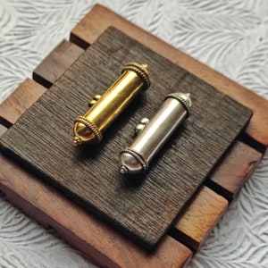 May include: Two small, cylindrical containers with decorative metalwork. One is gold and the other is silver. They are sitting on a wooden surface.