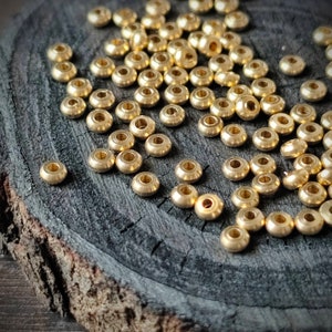 May include: A scattering of small, shiny, gold-coloured metal beads on a dark, textured wooden surface. Each bead has a rounded shape with a central hole, suitable for stringing. The beads are likely used for jewellery making or other crafts.