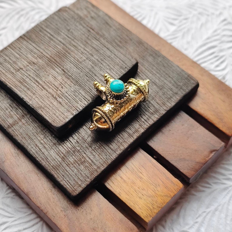 May include: A gold-colored pendant with a turquoise stone in the center. The pendant is shaped like a small cylinder with intricate details.