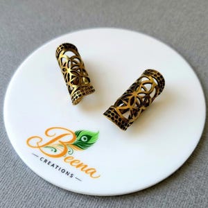 May include: Two cylindrical hair beads with an antique brass finish, featuring an intricate openwork geometric pattern of interlocking circles and a dotted border design at the top and bottom edges, displayed on a white circular surface.