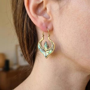 May include: A gold-toned hoop earring featuring a teardrop-shaped frame with an iridescent abalone shell inlay. The metal structure has a pointed, symmetrical design with a central marquise-shaped abalone piece and smaller accents.