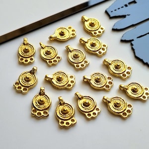 以下が含まれることがあります： 小さな金色の金属チャームのコレクション。各チャームは、中央に隆起した点と装飾的な三葉のベースを備えた円形のデザインが特徴です。チャームは白い表面に配置されています。