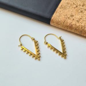 Può includere: Un paio di orecchini color oro. Ogni orecchino ha un design a forma di V con piccoli abbellimenti rotondi, distanziati uniformemente, lungo il bordo inferiore. Gli orecchini sono su una superficie bianca, con uno sfondo nero e color sughero.