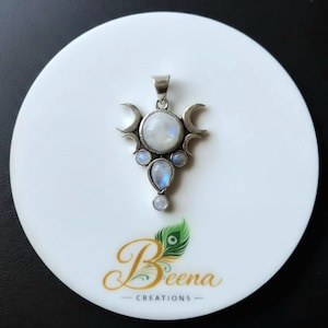 May include: A silver-toned pendant featuring a central round moonstone, a pear-shaped moonstone below, and two smaller round moonstones on the sides. The metal frame includes crescent moon motifs on both sides. The text Beena Creations is below.