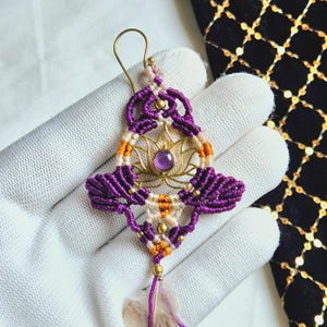 May include: Purple and orange macrame earrings with a gold lotus charm and a purple gemstone. The earrings have a feather tassel.