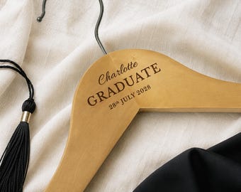 May include: A wooden clothes hanger with a metal hook. The hanger is engraved with the name "Charlotte", the word "GRADUATE", and the date "28th JULY 2028". A black tassel with gold accents is visible on the left.