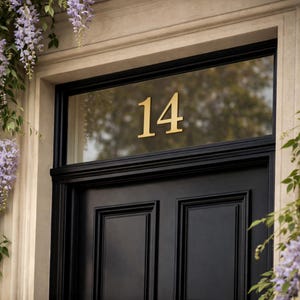 May include: A black front door featuring the number 14 in gold. The door is set within a black frame and a light stone surround. Wisteria flowers hang down the side of the door, adding a touch of colour.