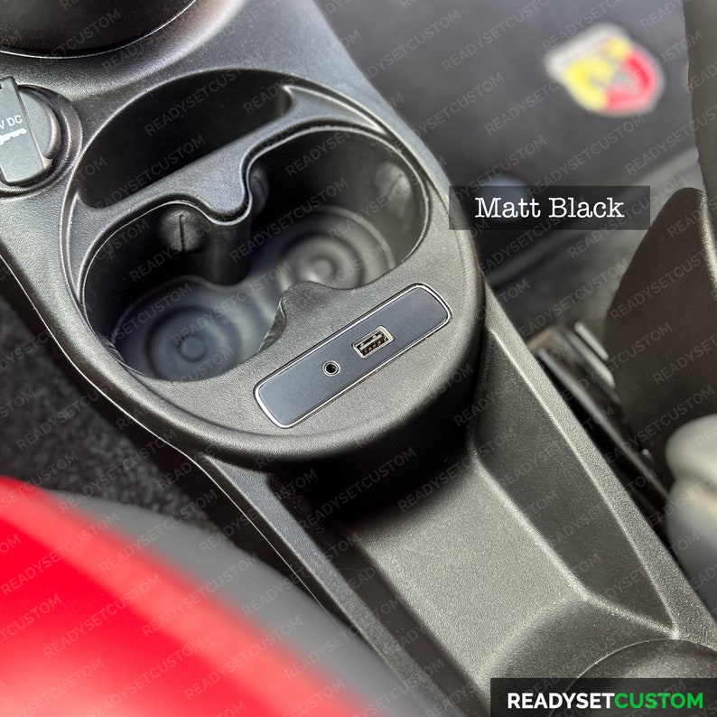 May include: A close-up of a black car interior with two cup holders and a USB port. The cup holders are empty and the USB port is covered with a black plastic cap. The interior is made of a textured material.